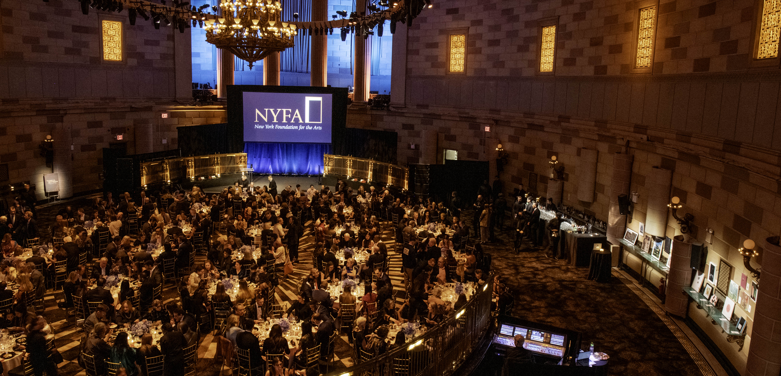 Aerial shot of NYFA's 2025 Hall of Fame Benefit at Gotham Hall in Manhattan, showing guests mingling at tables, some of NYFA's art auction, and a screen with the NYFA logo on it.