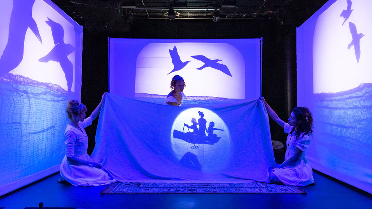 Three actresses dressed in white, who play sisters, are bathed in blue light while surrounded by three white screens also bathed in blue light. The the three screens have shadow puppetry ocean and seagulls flying. Two sisters in the middle the stage whole a blue sheet and the middle sisters lights a shadow puppet boat with three girls in it.