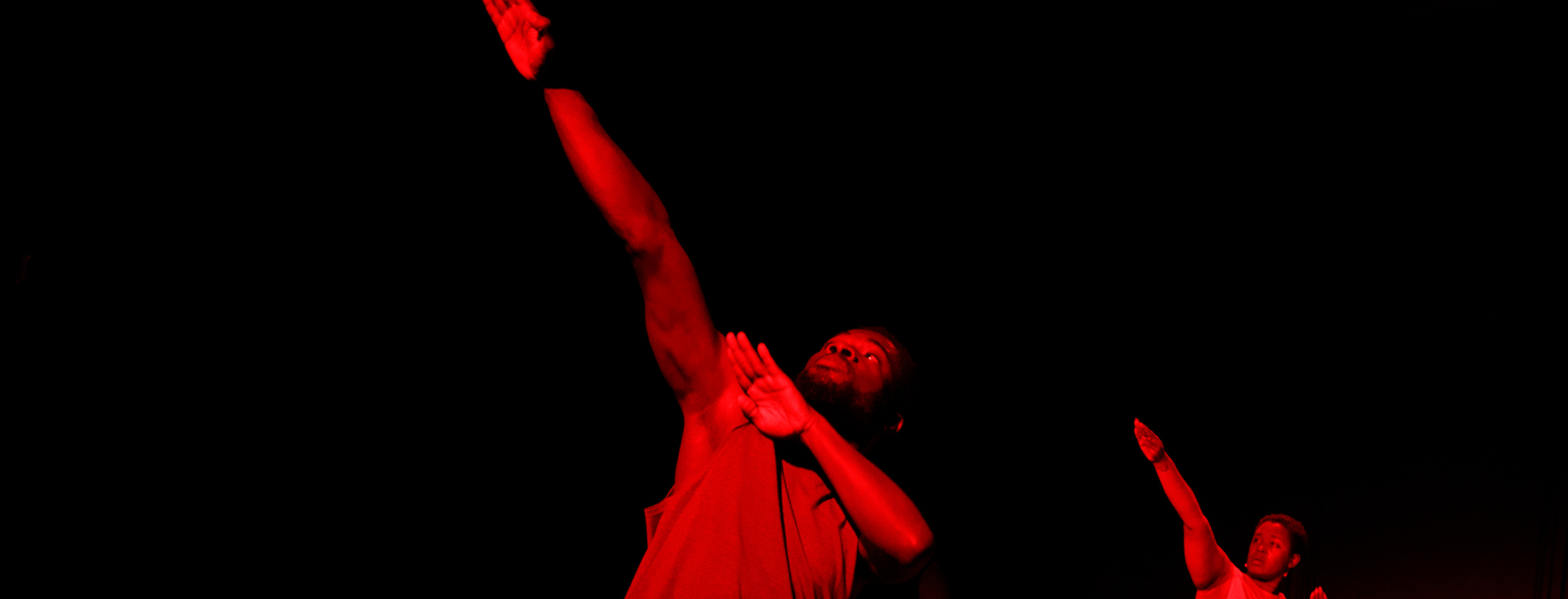 Cropped image of two dancers bathed in red light with one arm angled and raised against a black background.