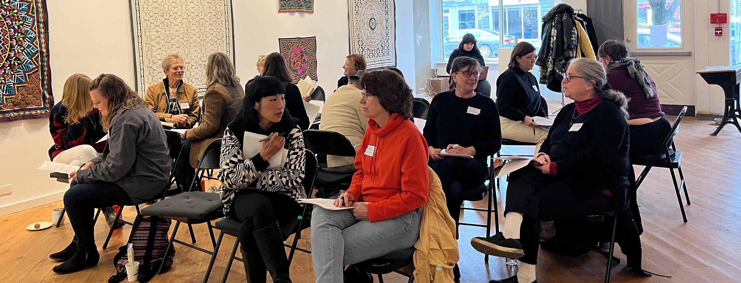 More than a dozen people are seated in an art gallery setting, grouped in twos and in conversation with one another