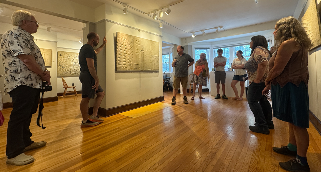A group stands in a semi-circle, their attention directed towards an individual who stands next to a flat, sculpture artwork hung on a wall in an exhibition space