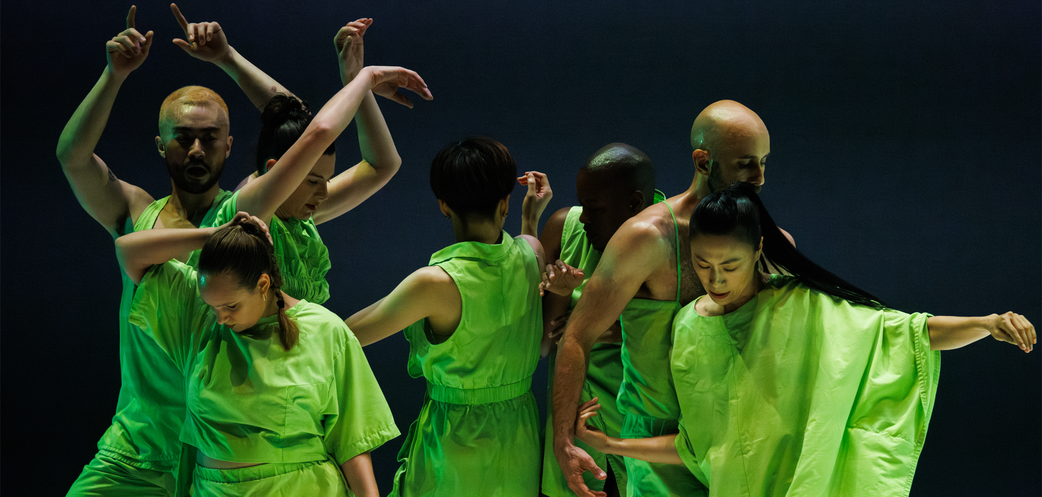 Seven dancers on stage wearing lime green standing in various poses.