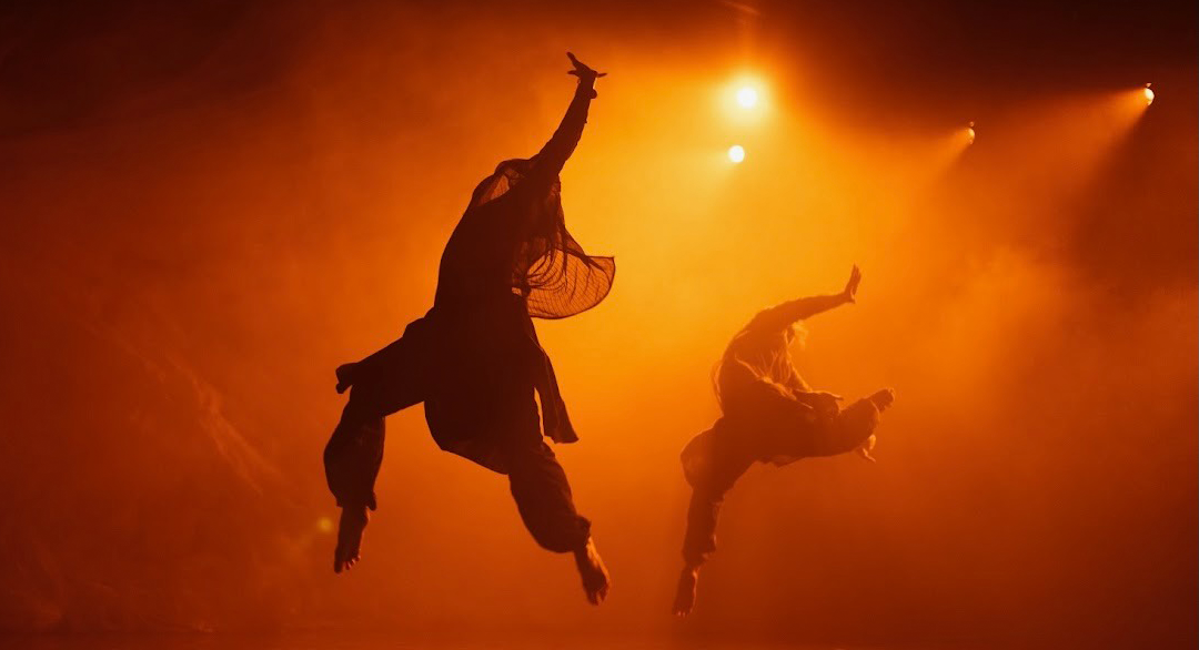 In burning orange lighting with haze in the air, two dancer flip jump in the air while turning from front to the back.