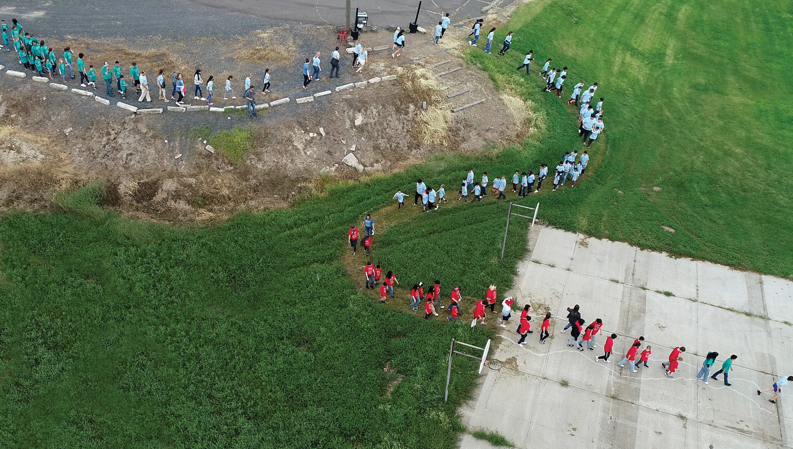 Aerial drone shot of school children of varying ages walk in pairs in a curving line in the shape of a river. The students wear matching t-shirts in turquoise, sky blue, or red that bear the project's logo.