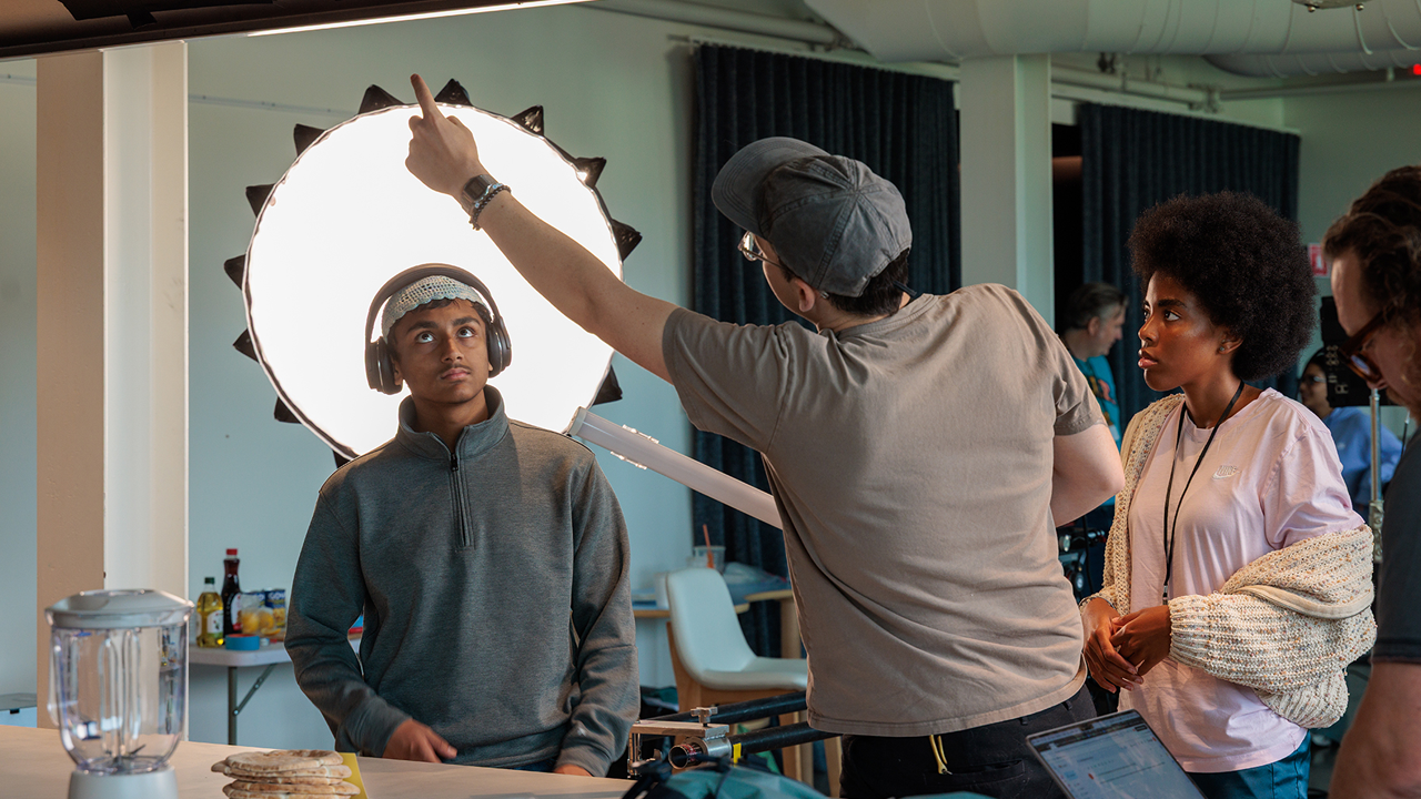 An instructor, stands in a classroom with his back to the camera and holds an LED strip light in one hand while pointing up with the other; two teen students facing the camera look up to where the instructor is pointing, one of whom stands in front of brightly lit light dome.