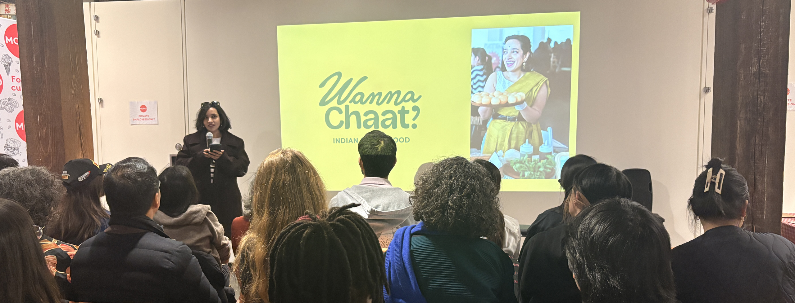 An individual stands before group of people giving a presentation, microphone in hand. Projected is a bright yellow graphic with the words 