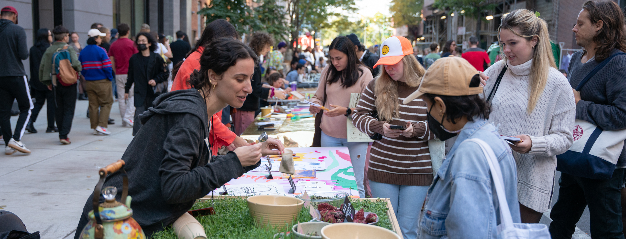 People stand around a table with a tablecloth made of living sprouts, a woman is talking and showing other people herbs in bowls that sit on the table.