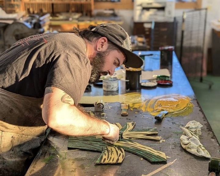 A bearded artist leans over a workbench in a printshop. Using a rubber brayer, he is rolling green and yellow ink onto a hand-carved masonite block.