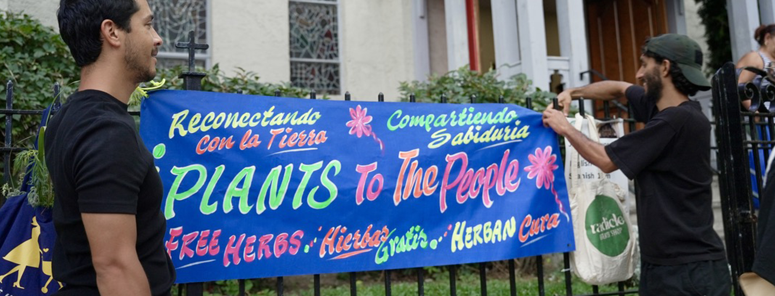 One person walks by as another affixes a "Plants to the People" sign to a metal gate. The sign features words in English and Spanish that advertise free herbs and reconnecting with the earth.