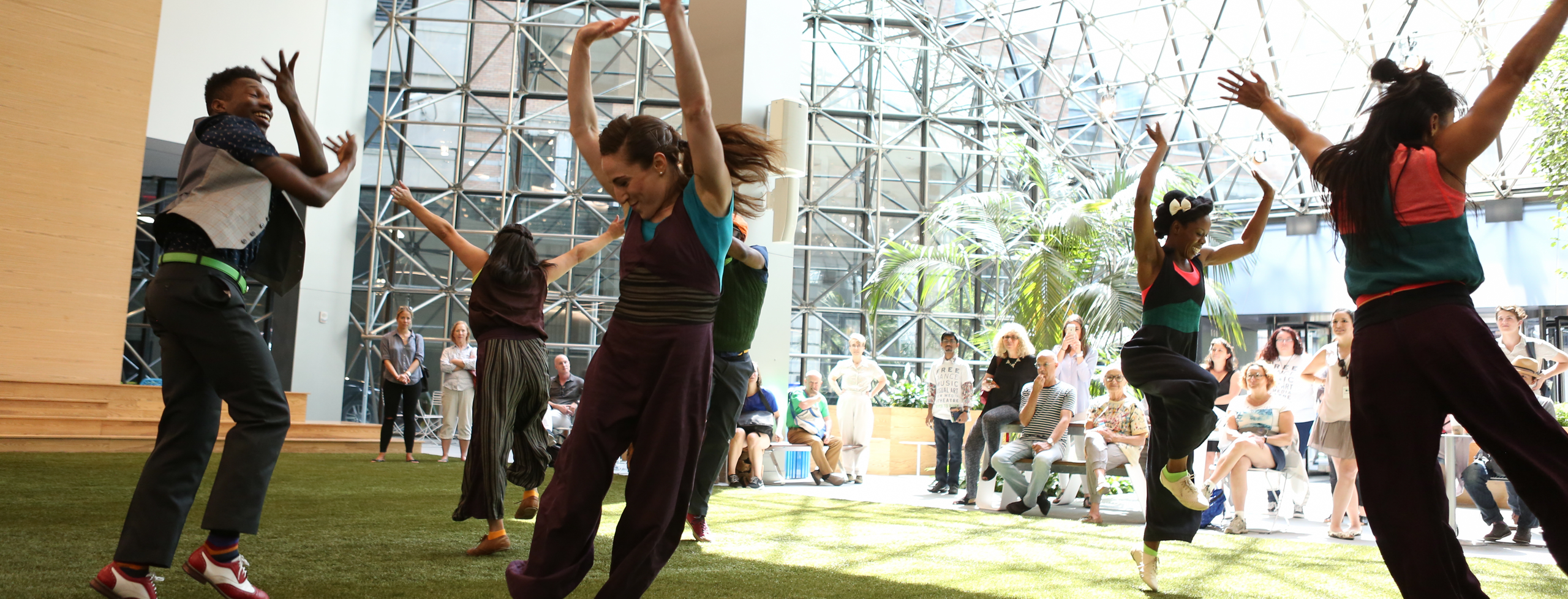 Five dancers wearing outfits that are navy blue, grey, dark purple and green throw their arms up in the air at various angles. They create a circle together and are md-step; their energy is celebratory and uplifting. They are dancing on the green astro-turf of a public atrium with beams of sunshine streaming though the glass windows surrounding them.