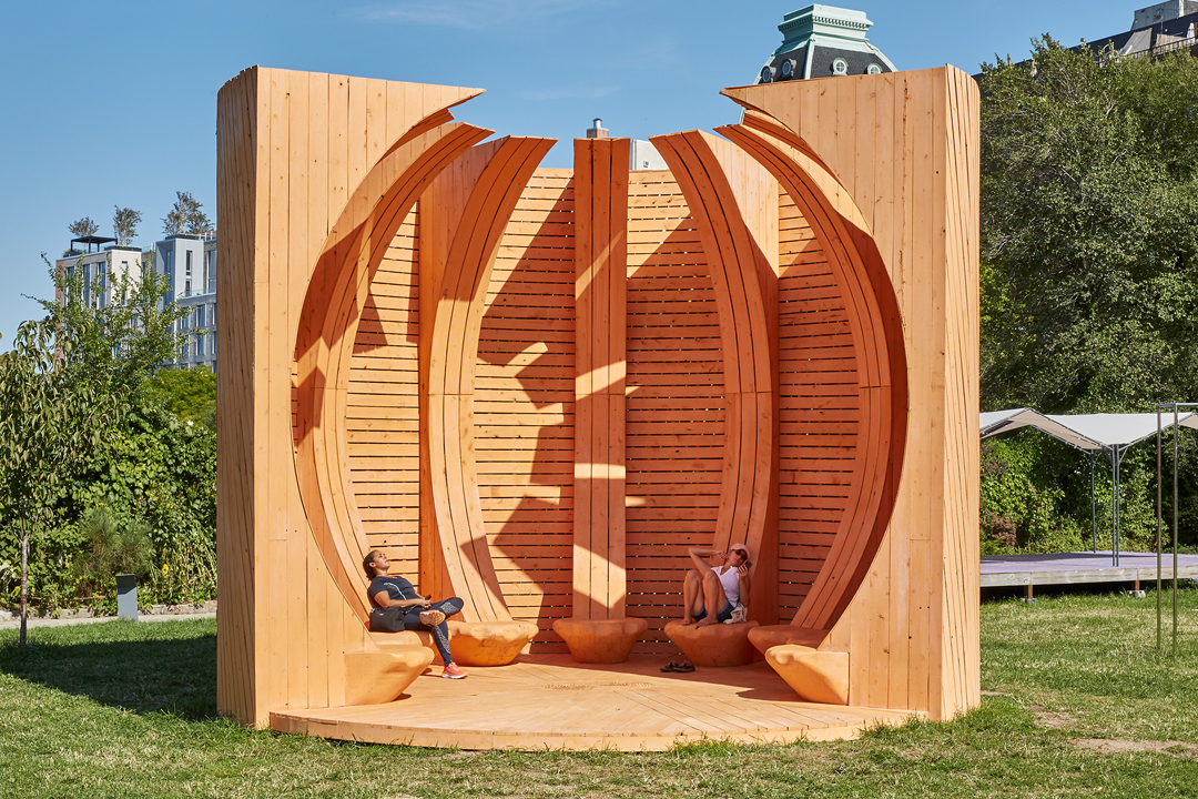 striking wooden pavilion structure set outdoors on a grassy area. The design resembles a partially open coral polyp, composed of tall vertical wooden slats and curved ribs that arch overhead.
